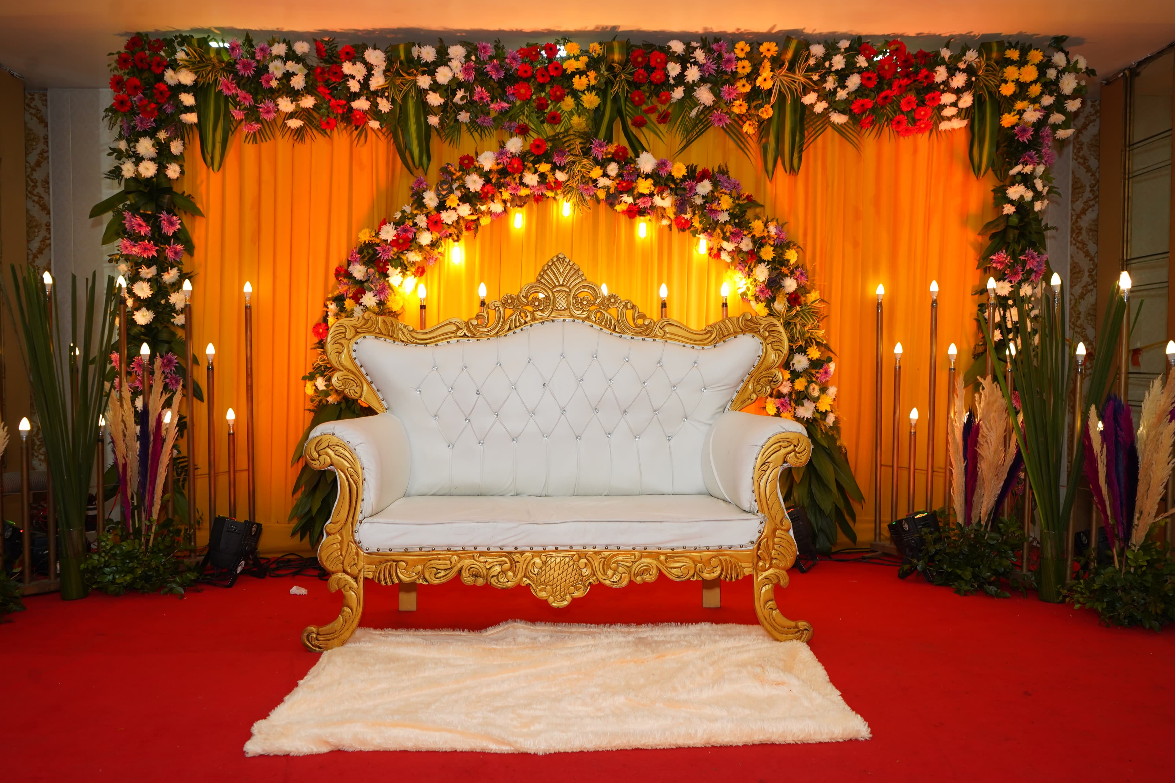 Ornate wedding stage with gold sofa, floral arch, and warm candle lighting